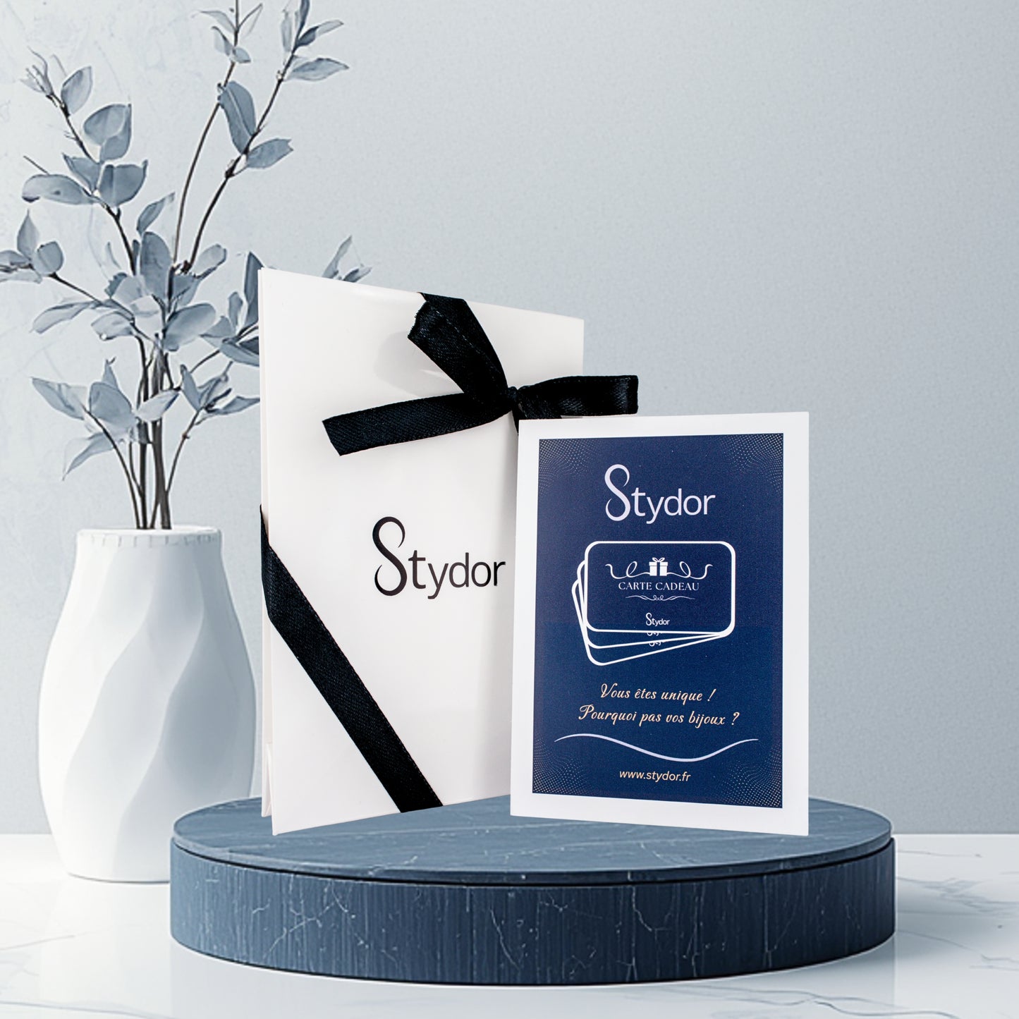 Visuel du coffret carte cadeau Stydor : pochette fermée par un ruban noir avec une carte cadeau à côté, envoyé par voie postale, valable un an, offrant un bijou personnalisé sur mesure, cadeau prestigieux et chargé d’émotion.